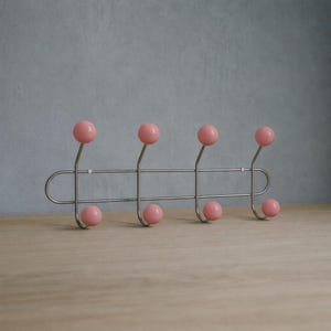 May include: A retro-style wall-mounted coat rack with a polished chrome finish. It features six pink ball-shaped hooks, three on top and three below, arranged in a horizontal line. The rack is mounted on a light wooden surface.