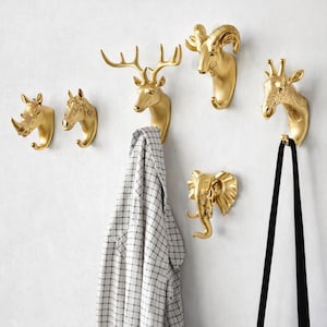 Puede incluir: Un conjunto de ganchos de pared con cabeza de animal dorada, que incluye un rinoceronte, un caballo, un ciervo, un carnero, una jirafa y un elefante. Una camisa a cuadros y una correa negra cuelgan de los ganchos. Los ganchos están montados en una pared blanca.