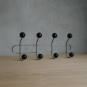 Könnte beinhalten: Ein moderner, an der Wand montierter Kleiderhaken mit sechs schwarzen, kugelförmigen Knöpfen. Der Haken hat eine gebürstete Metalloberfläche und ein horizontales Stangendesign. Die Knöpfe sind gleichmäßig verteilt. Auf einer hellen Holzoberfläche montiert.