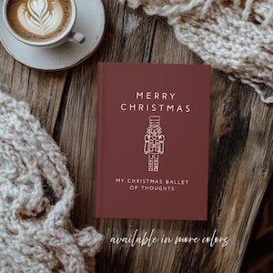 Puede incluir: Un cuaderno burdeos con el texto "MERRY CHRISTMAS" y una ilustración blanca de un cascanueces. El cuaderno también tiene el texto "MY CHRISTMAS BALLET OF THOUGHTS" y "available in more colors". Una taza de café y una bufanda están en el fondo.