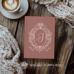 May include: A dusty rose-colored journal with a white ornate design featuring the letter "J" inside an oval frame. The journal is on a wooden surface, with a cup of coffee and a knitted scarf nearby. Text at the bottom reads "available in more colors."