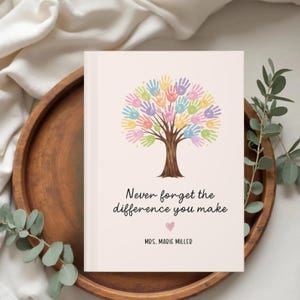 Puede incluir: Un diario rosa claro con un diseño de árbol hecho de huellas de manos coloridas. El texto dice "Nunca olvides la diferencia que haces" con un corazón y el nombre "Mrs. Marie Miller".