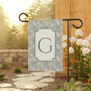 May include: A garden flag with a floral design and a large letter "G" in a framed shape. The flag is displayed on a black metal stand in a garden, with flowers and greenery in the background, creating a decorative outdoor accent.