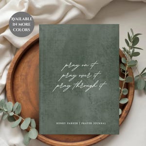 Könnte beinhalten: Ein grünes Gebetbuch mit weißer Schrift, die "pray on it, pray over it, pray through it" lautet. Das Buch liegt auf einem Holztablett mit Grünzeug. Der Text "HENRY PARKER | PRAYER JOURNAL" steht unten.
