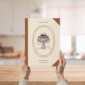 May include: A recipe journal with a cream-colored cover and a brown spine. The cover features a cake illustration inside a decorative oval frame. The text "MICHELLE SAMUELS" is at the top, and "RECIPE JOURNAL" and "from fridge to plate" are at the bottom.