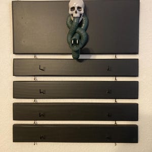 May include: A wall-mounted organizer featuring five horizontal black rectangular panels connected by metal links. The top panel displays a three-dimensional white skull with a dark green snake coiled around it, while the lower four panels have small black hooks.