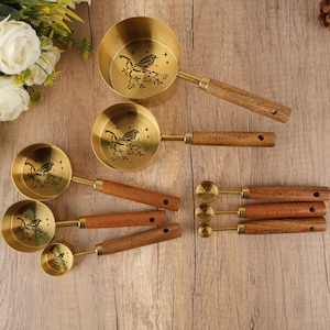 May include: A collection of brass measuring cups and spoons with wooden handles. Each cup and spoon has a bird and branch design. The handles are a dark brown colour. The largest cup is labelled "1/2 cup / 125ml".