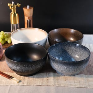 以下が含まれることがあります： 黒、茶色、クリーム色、青灰色の4つのセラミックボウル。斑点模様があります。ボウルは模様入りのプレースマットの上に配置されています。金色のボトルと木製のペッパーグラインダーが背景にあります。