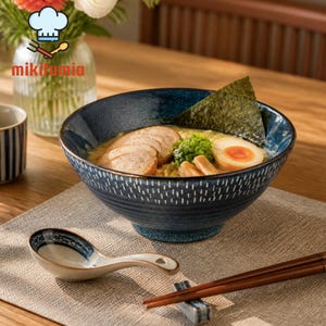 以下が含まれることがあります： 麺、豚肉のスライス、ゆで卵、海苔が入った青と白の模様のラーメン丼。お揃いのスプーンと箸が、ニュートラルカラーのプレースマットの上に置かれています。丼には金の縁取りがあり、「mikifumia」の文字が入っています。