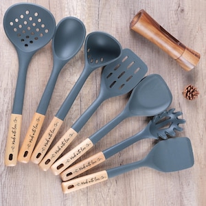 以下が含まれることがあります： 木製の柄が付いたグレーのシリコン製キッチン用品のセット。セットには、穴あきスプーン、無垢のスプーン、おたま、穴あきヘラ、無垢のヘラ、パスタサーバー、木製ペッパーグラインダーが含まれます。ハンドルには「Made with love」と刻印されています。