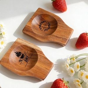 以下が含まれることがあります： 天然木目の2つの木製スプーンレスト。1つはグリルグラフィックと下に「AMY」という名前があります。もう1つは「Pumpkin＆Popcorn」というテキストが入ったリースデザインです。イチゴとデイジーが散らばっています。