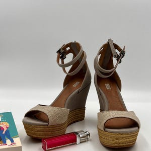 Könnte beinhalten: Paar beige Keilsandalen mit Knöchelriemen und geflochtenen Plateaus. Die Sandalen haben offene Zehen und werden mit einem roten Lippenstift und einem kleinen Buch präsentiert.