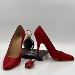 Könnte beinhalten: Ein Paar rote High Heels mit strukturiertem Muster. Die Schuhe werden mit einer Parfümflasche, einem Buch und einer Flasche rotem Nagellack präsentiert. Die Schuhe haben eine spitze Zehenpartie und einen Stiletto-Absatz.