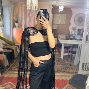 以下が含まれることがあります： 黒いフリンジショール、ストラップレスのトップス、黒いパンツ。ショールは長いフリンジが特徴的なユニークなデザインです。この服装は、黒いレザー風パンツで完成しています。背景にはカラフルなラグと鏡があります。