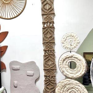 May include: A long, vertical macrame wall hanging made of tan cord with intricate geometric knot patterns, suspended from a wooden dowel. Beside it are three stacked circular white wool fiber art pieces and a light gray, irregular shaped object.