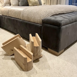 May include: Wooden bed risers in a natural finish are displayed in front of a gray upholstered bed with a matching storage bench. The bed is covered with a beige knitted throw and pillows.