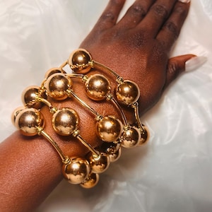 May include: Gold-coloured bangles featuring large, spherical beads. The bracelets are worn on a dark-skinned arm, with long, pointed fingernails. The jewellery is set against a white, textured background. The bangles are a statement piece.