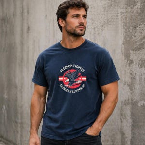 May include: Dark blue t-shirt with a circular emblem depicting an eagle, red and white stripes, and the text "FREEDOM FIGHTER" and "AMERICAN AUTHENTIC". The shirt has short sleeves and a round neck.