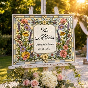 May include: A rectangular wedding sign featuring a stained glass style border with colourful pink, yellow, and purple flowers. The centre panel displays the text The Millers, Olivia & Adams, and the date 05.20.2027 in a black script and serif font.