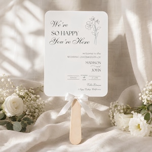 May include: A white rectangular hand fan with rounded corners featuring black text that reads We're SO HAPPY You're Here, wedding celebration of Madison and John, September 28, 2028, at 3 PM, Villa Eterna, Napa Valley, California, and a white ribbon.