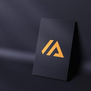 May include: A black business card with a gold logo featuring the letters "IA" in a stylized design. The card is leaning against a dark surface, with a shadow cast across the background.