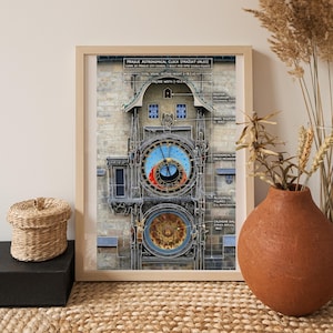 May include: Framed print of the Prague Astronomical Clock (Pražský Orloj). The clock face features a blue background with gold accents and intricate details. The print includes architectural details and text labels. The frame is a light wood color.