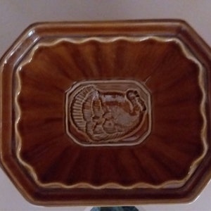 May include: A brown ceramic dish with a raised design of a turkey in the center. The dish has a scalloped edge and a slightly octagonal shape.