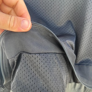 May include: Close-up of a dark gray jacket with a mesh lining. The jacket has a zipper closure and a leather-like trim at the bottom. The mesh lining is visible through the open zipper.