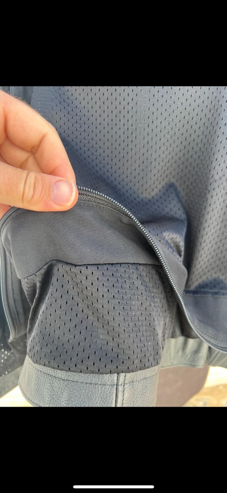 May include: Close-up of a dark gray jacket with a mesh lining. The jacket has a zipper closure and a leather-like trim at the bottom. The mesh lining is visible through the open zipper.