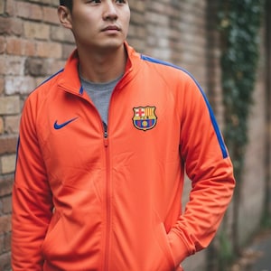 Könnte beinhalten: Orangefarbene Trainingsjacke mit blauem Akzent an den Schultern und einem blauen Nike-Logo. Die Jacke hat ein Teamwappen auf der Brust. Die Person trägt ein graues T-Shirt und blaue Jeans.