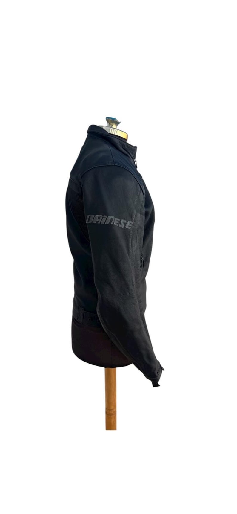 May include: Black motorcycle jacket with the brand name "DAINESE" printed on the left sleeve. The jacket is displayed on a mannequin. The jacket has a high collar and a zipper closure. The jacket is made of a durable material.