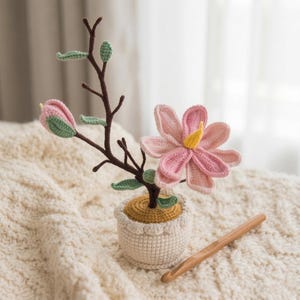 Op de afbeelding: Een gehaakte bloemstuk met een roze bloem en een gele kern, een roze knop en groene bladeren. De opstelling staat in een kleine, gebroken witte pot met een gele basis. Een houten haaknaald ligt in de buurt.