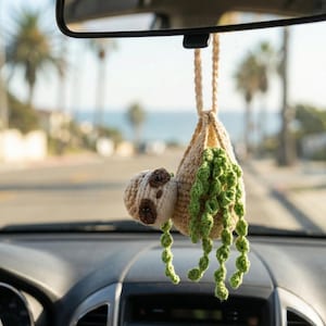 May include: A crocheted sloth car charm suspended from a car's rearview mirror. The sloth is beige with brown markings and button eyes. It hangs from a beige pouch with green, vine-like strands. The background shows a sunny street and the sea.
