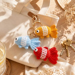 May include: Three crochet goldfish keychains in yellow, blue, and red, each with white accents. The keychains are attached to gold-colored metal rings and clasps. A beige tote bag is in the background.
