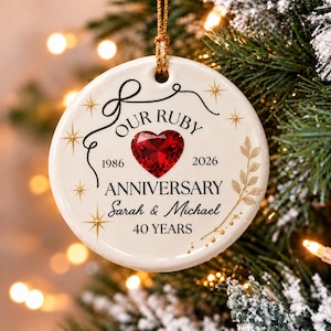 Op de afbeelding: Een rond wit keramieken ornament met een gouden ophangkoord. Het bevat een rood hart, de tekst "OUR RUBY ANNIVERSARY", "1986", "2026", "ANNIVERSARY", "Sarah & Michael" en "40 YEARS", versierd met gouden sterren en een takje met blaadjes.