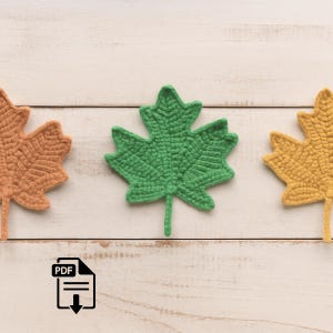 Puede incluir: Tres hojas de arce de ganchillo en tonos naranja, verde y amarillo están dispuestas sobre una superficie de madera blanca. Las hojas están detalladas con patrones de puntadas visibles, lo que sugiere una artesanía hecha a mano. La imagen evoca un tema otoñal.