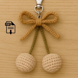 May include: A handmade crochet keychain in the shape of cherries. The keychain features a tan bow, green stems, and two beige spheres. The keychain is attached to a silver metal clasp. The background is a light brown wooden surface.