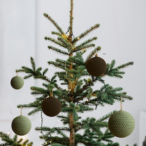 Op de afbeelding: Een kerstboom versierd met verschillende handgemaakte gehaakte ornamenten in groen- en bruintinten. De ronde ornamenten hebben een getextureerd uiterlijk en hangen aan de takken van de boom. De boom is verlicht met kleine witte lichtjes.