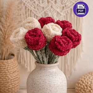 Op de afbeelding: Een boeket handgemaakte gehaakte bloemen in een gevlekte keramische vaas. De bloemen zijn een mix van crème en dieprood, met groene stelen. De achtergrond bevat een geweven wandkleed en een PDF-pictogram.