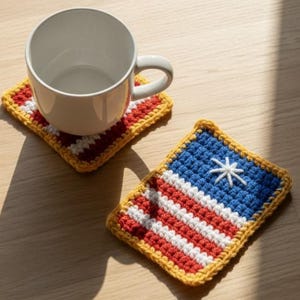 Op de afbeelding: Een witte keramische mok staat op een vierkante, gehaakte onderzetter met een rood-wit-blauw vlagontwerp. Een tweede onderzetter met hetzelfde ontwerp staat ernaast. De onderzetters hebben een gele rand.