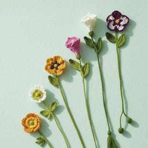 以下が含まれることがあります： 白、黄色、ピンク、紫など、さまざまな色の手作りのかぎ針編みの花のブックマークのコレクション。各ブックマークは、詳細な花の頭、緑の葉、長い緑の茎が特徴です。ブックマークは、水色の背景に対して斜めに配置されています。