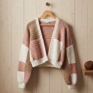 May include: A cropped, open-front cardigan in a patchwork design. The sweater features blocks of white, light pink, and tan colours. It hangs on a wooden hanger against a light wood-panelled wall. Two balls of yarn sit on a small shelf.