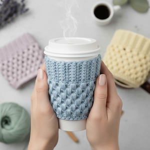 May include: A light blue knitted coffee cup sleeve with a textured pattern, held in hands. Steam rises from the white-lidded cup. Additional knitted sleeves in lavender and yellow are visible, along with a ball of green yarn.