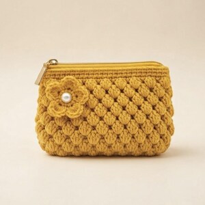 May include: A mustard yellow crochet pouch featuring a textured bobble stitch pattern. The rectangular accessory includes a gold-toned metal zipper closure and a decorative crocheted flower accent on the front, centered with a single white pearl bead.
