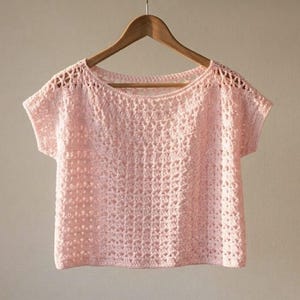May include: A light pink crochet top featuring a wide boat neckline and short sleeves. The garment is crafted with an openwork stitch pattern that creates a repeating grid-like texture throughout the body, hanging on a wooden clothes hanger.