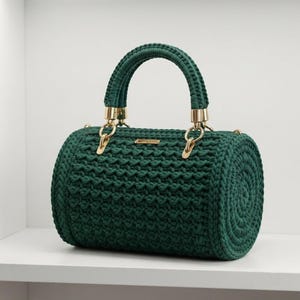 Puede incluir: Un bolso de mano cilíndrico de color verde oscuro hecho de cordón grueso tejido a ganchillo. El bolso cuenta con un asa tejida a juego, herrajes de metal dorado que conectan el asa al cuerpo y una pequeña placa rectangular dorada en el frente.