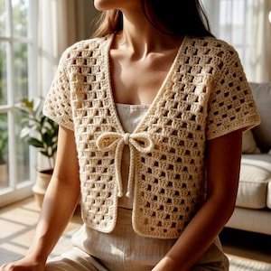 May include: Cream-colored crochet cardigan with short sleeves and a tie closure. The openwork design of the cardigan is visible, and the garment appears to be handmade. The cardigan is a light, neutral color, suitable for layering.