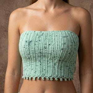 May include: A light mint green strapless tube top featuring a vertical ribbed crochet pattern with dark blue speckled accents throughout the fabric. The garment has a scalloped hemline with small loops along the bottom edge and a small side tag.