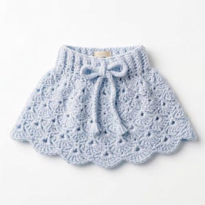 Op de afbeelding: Een lichtblauwe, gehaakte babycape met een geschulpte rand. De cape heeft een strik bij de halslijn met twee kwastjes. Het haakpatroon is open en luchtig en de cape is ontworpen om om de nek te worden gedragen.