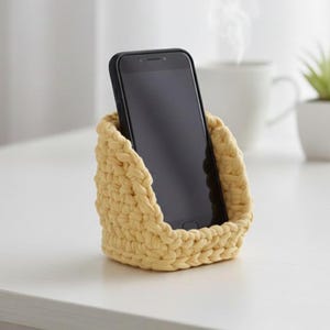 以下が含まれることがあります： 黒いスマートフォンが入った、薄い黄色の手編みの電話ホルダー。ホルダーは太い糸でできており、バスケットのようなデザインです。電話は直立しており、ホルダーは白い表面にあります。